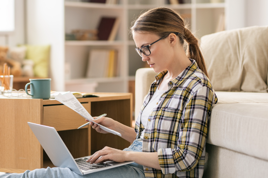 young woman sitting on floor in living room and using laptop to review her budget 